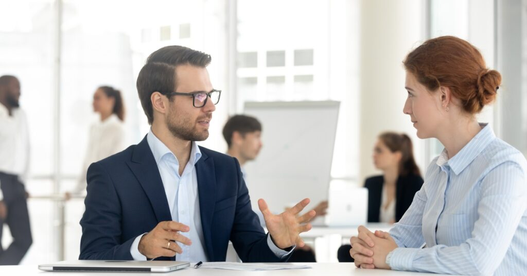 A man in a blue suit and glasses is talking to a woman in business attire at a white desk in an office.