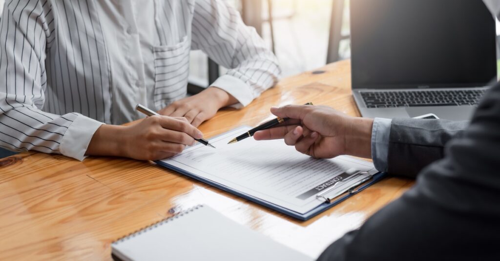 An over-the-shoulder view of a man in a suit instructing a woman at a table where to sign a contract with a pen.