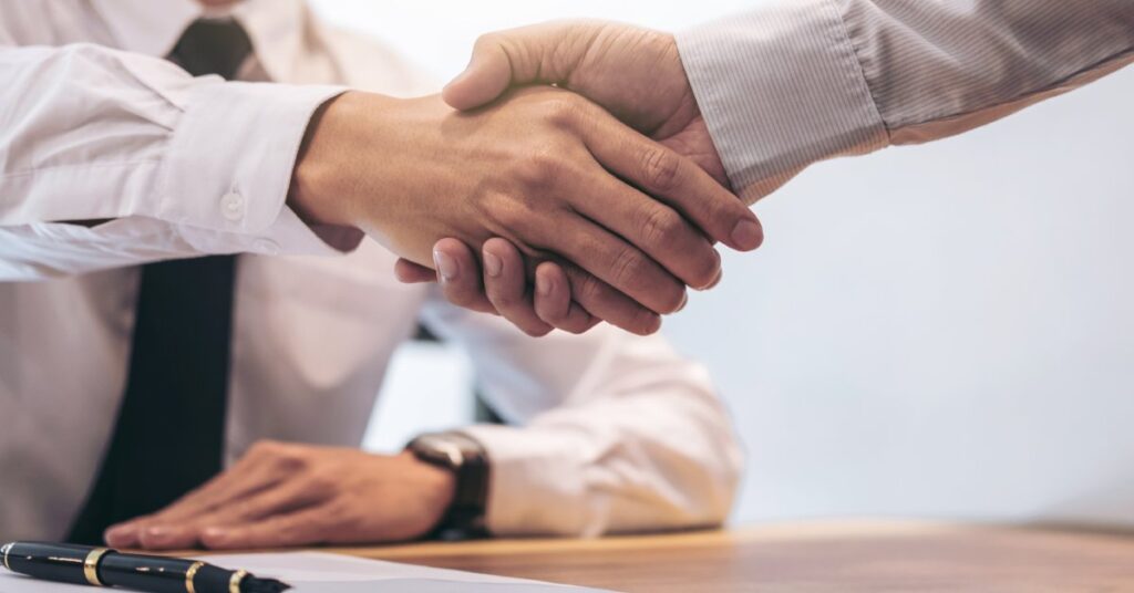 A close-up of two men in business attire shaking hands over a wooden table with a clipboard, documents, and a pen.