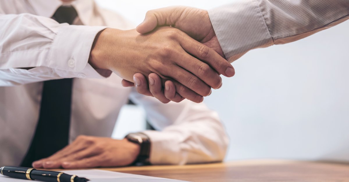 A close-up of two men in business attire shaking hands over a wooden table with a clipboard, documents, and a pen.