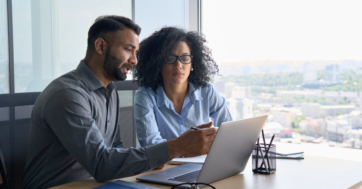 A man and woman, both in business attire, sitting next to and talking to one another at a desk in an office with a view.
