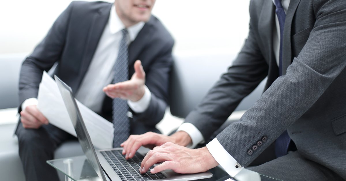 A close-up of two men in suits sitting on couches and talking to one another. One is typing on a laptop computer.