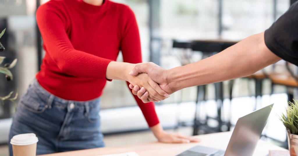 A woman in a red sweater and jeans smiles as she shakes the hand of someone over a desk with a laptop and business papers.