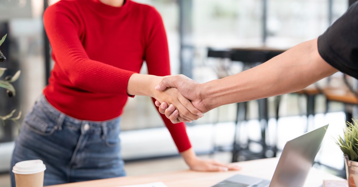A woman in a red sweater and jeans smiles as she shakes the hand of someone over a desk with a laptop and business papers.