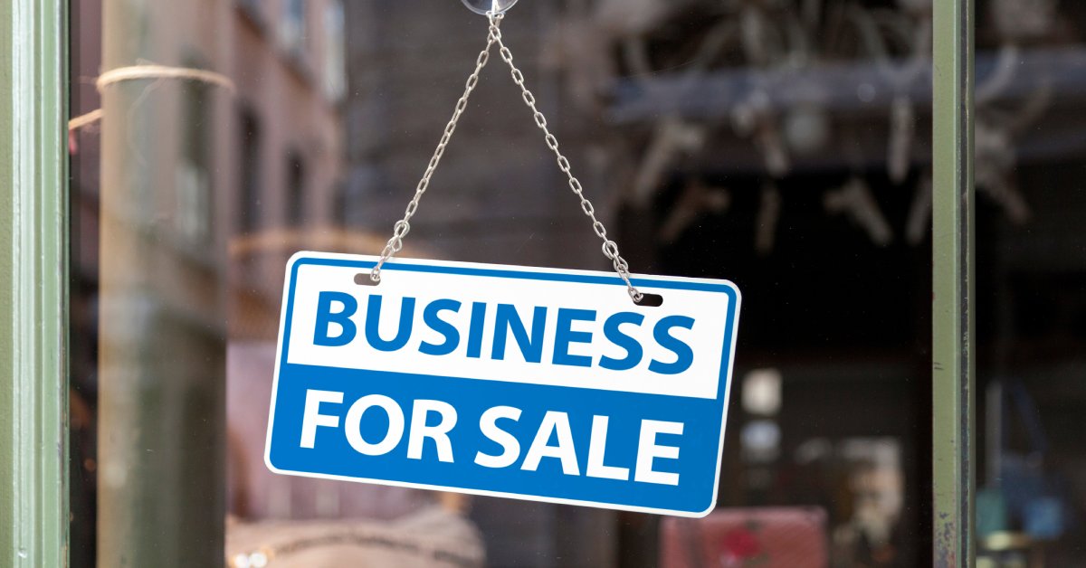 A small, blue and white sign hanging from a suction cup and chain in a store window that reads "BUSINESS FOR SALE."