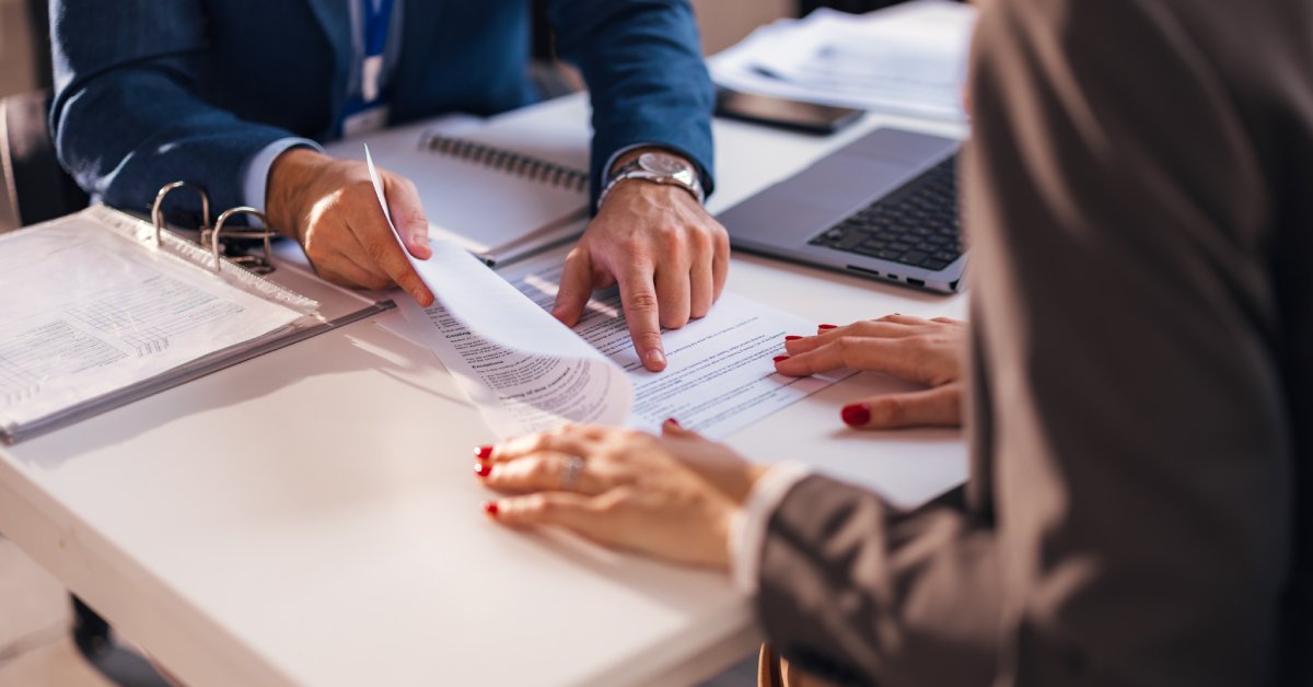 A man in a business suit in front of various papers and binders pointing to text on a document for a woman.
