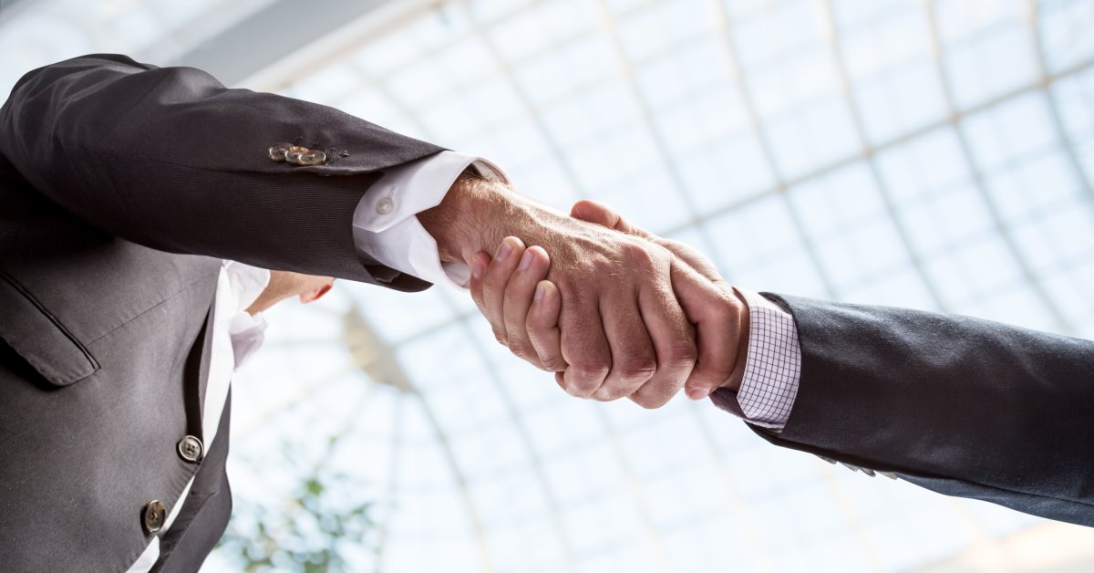 An underneath view of two men in business suits shaking hands. Above the two men is a glass skylight ceiling.