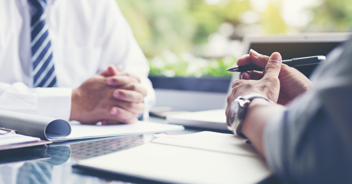 A close-up of the clasped hands of two people while they sit at a desk with papers in front of them and discuss.