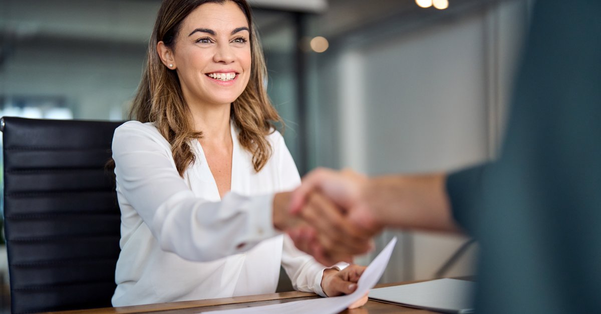A woman in a white blouse sitting in a desk chair smiles as she reaches across the desk to shake someone's hand.