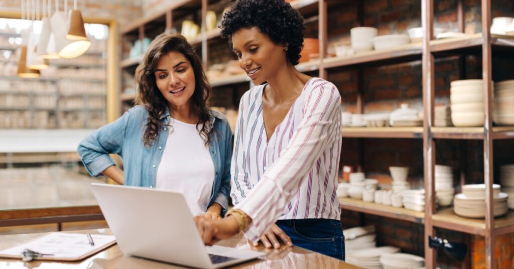 Two women in a ceramic workshop discuss while pointing to a laptop open on a table in front of them.