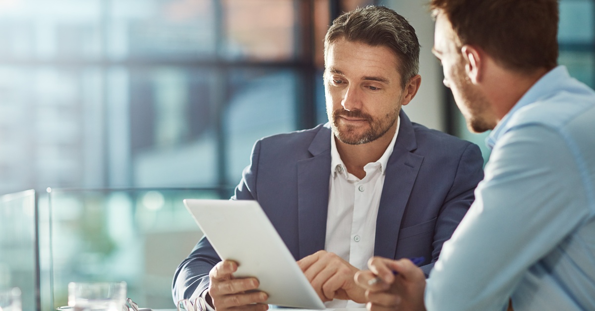 A man in a blue business coat and white shirt examines an electronic tablet next to another man at a white table.