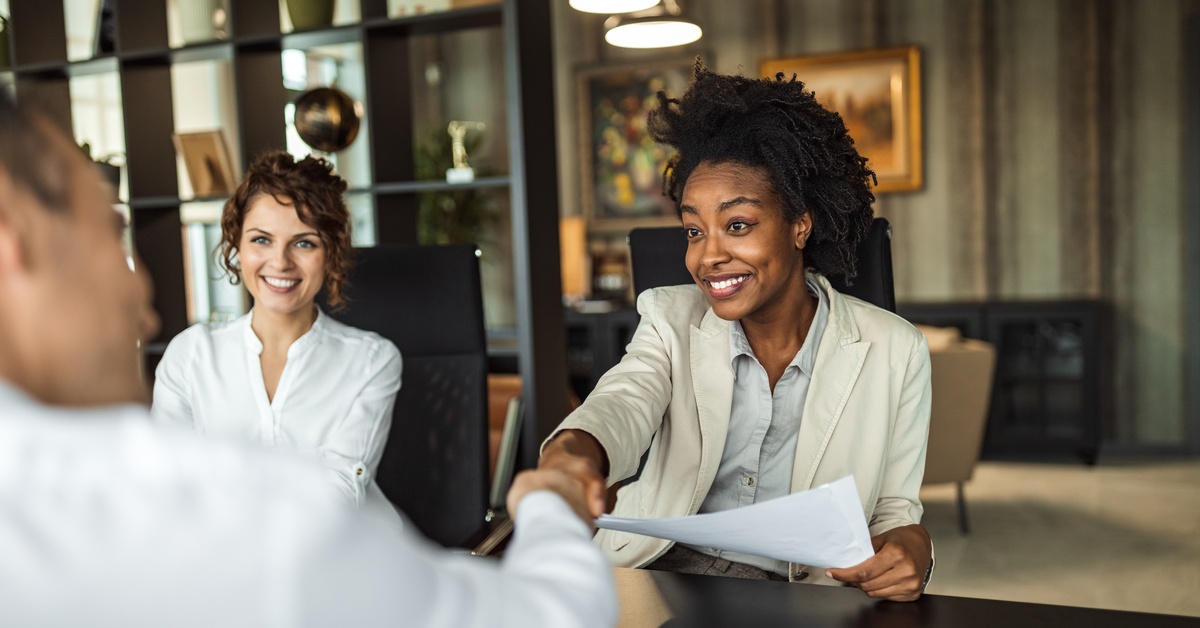 A woman holding papers reaches across a table to shake hands with a man, with another woman sitting next to her.