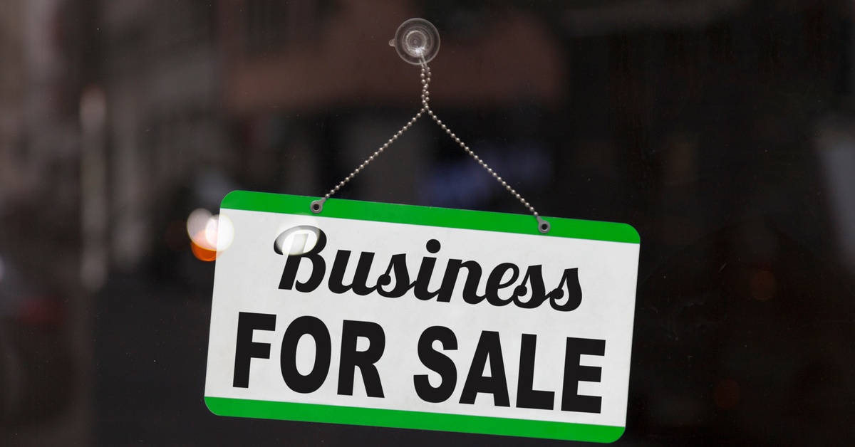 A green and white business sign hanging from a chain in a glass window with "Business FOR SALE" in black text.