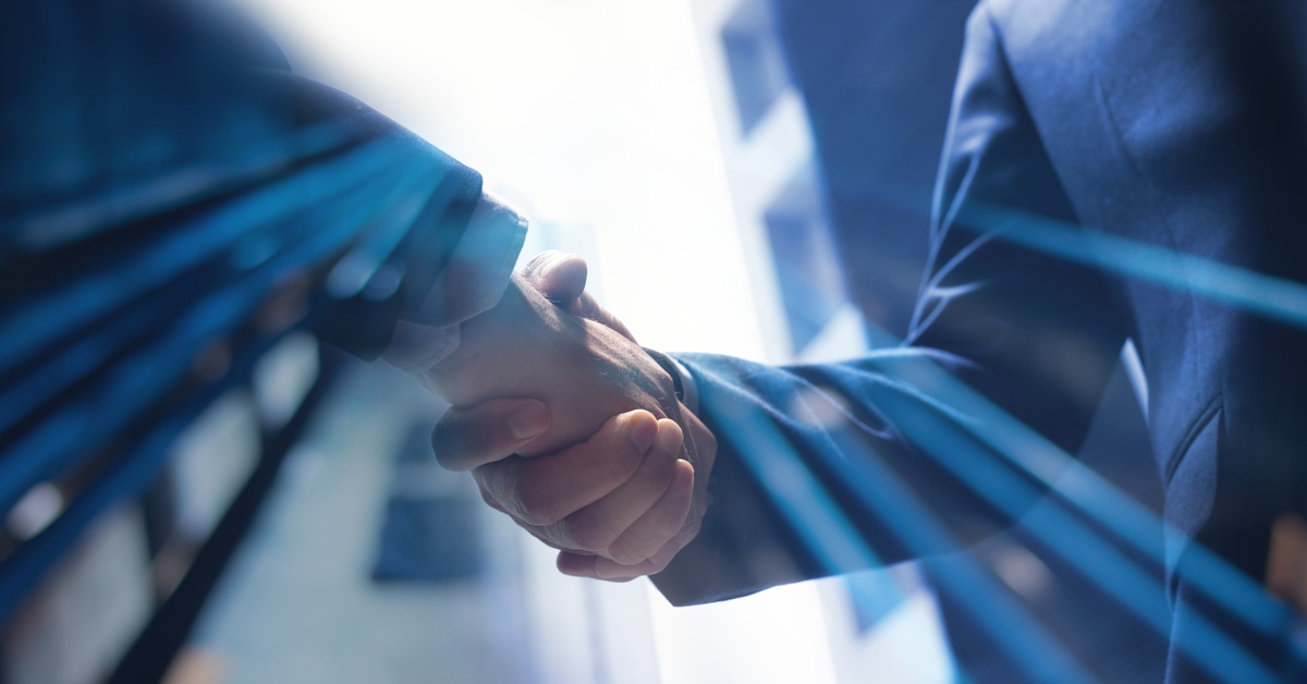 A close-up of two men in business suits shaking hands on a sunny day with buildings in the background.