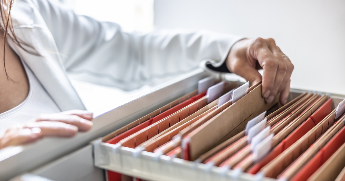 A close-up of a woman in a white shirt looking through a file cabinet and pulling out a labeled folder.