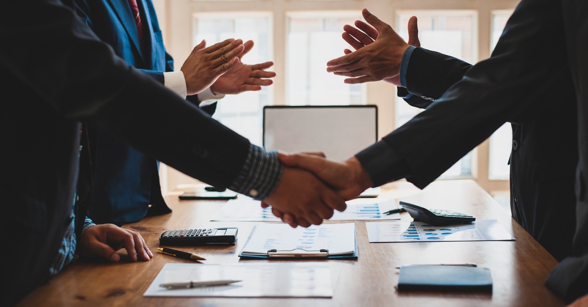 Two people in business suits shaking hands over a wooden table with various documents as two other people applaud.