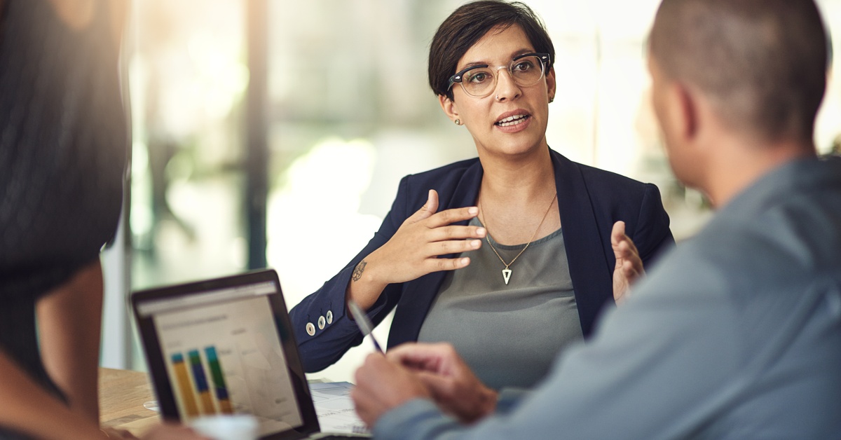 A woman talks to a person sitting at a wooden table with a laptop in front of them, with statistical graphs.