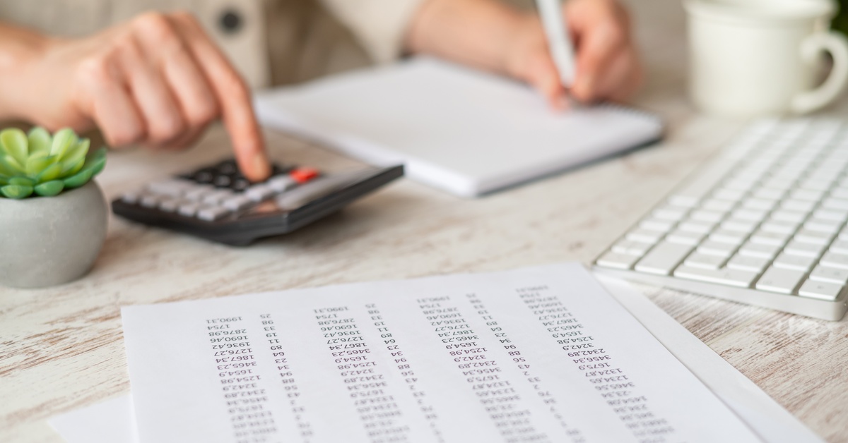 A close-up of a woman at a desk using a calculator and writing in a notebook in front of a keyboard and document.