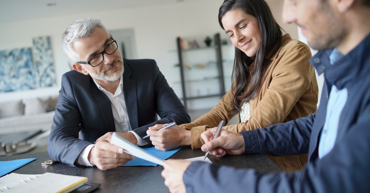 An older man with a white beard and glasses in a suit watches as a man and a woman sign documents at a table.