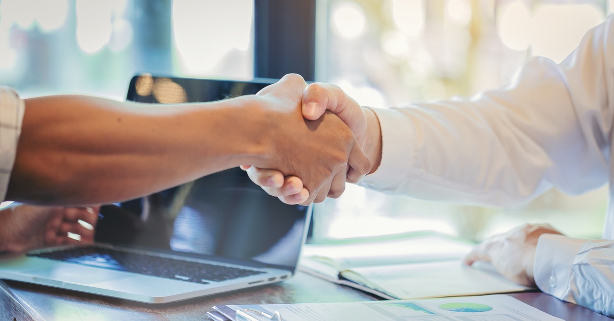 A close-up of two men shaking hands over a wooden table with a laptop, clipboards, and booklets on it.