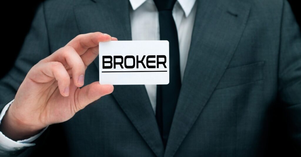 A close-up of a man in a suit, with a white shirt and black tie, holding a white business card that says "BROKER."