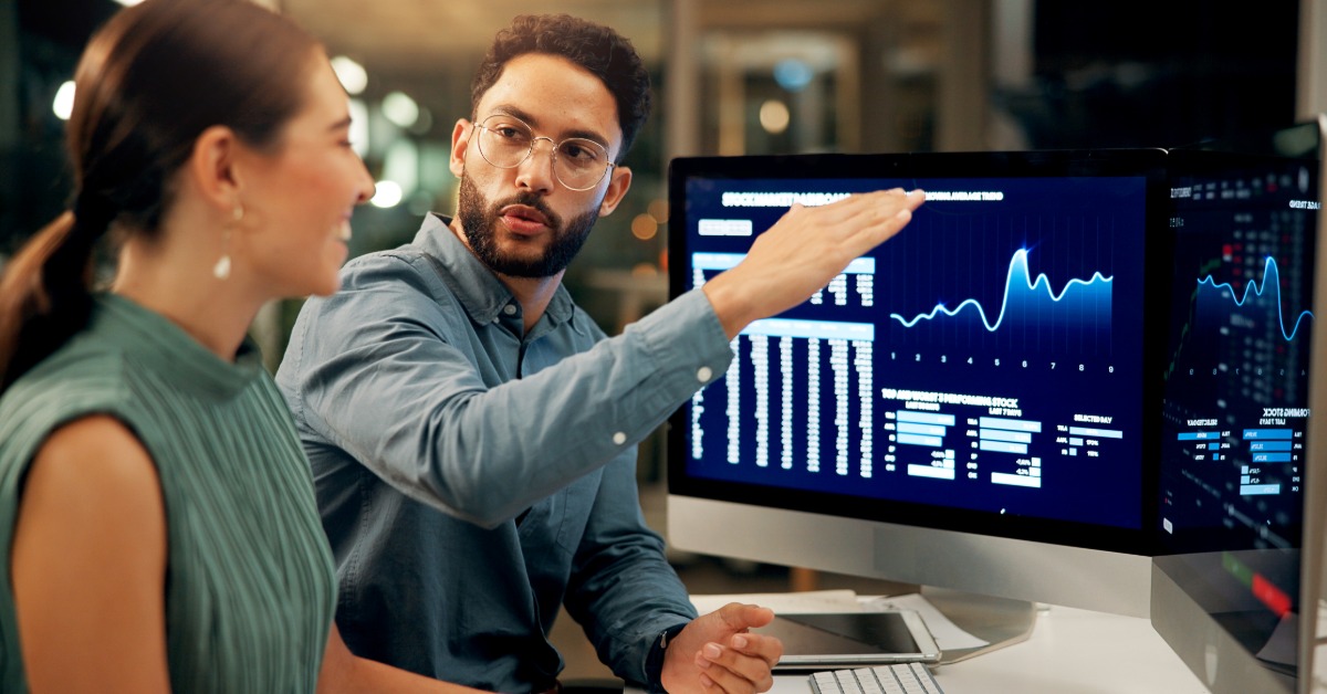 A man with glasses and a beard explaining something to a woman while sitting at a desk with multiple desktop displays.