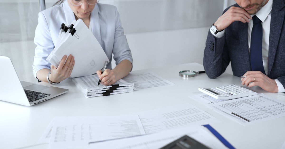 A man and a woman in business attire are sitting at a white desk, intently looking at stacks of financial documents.