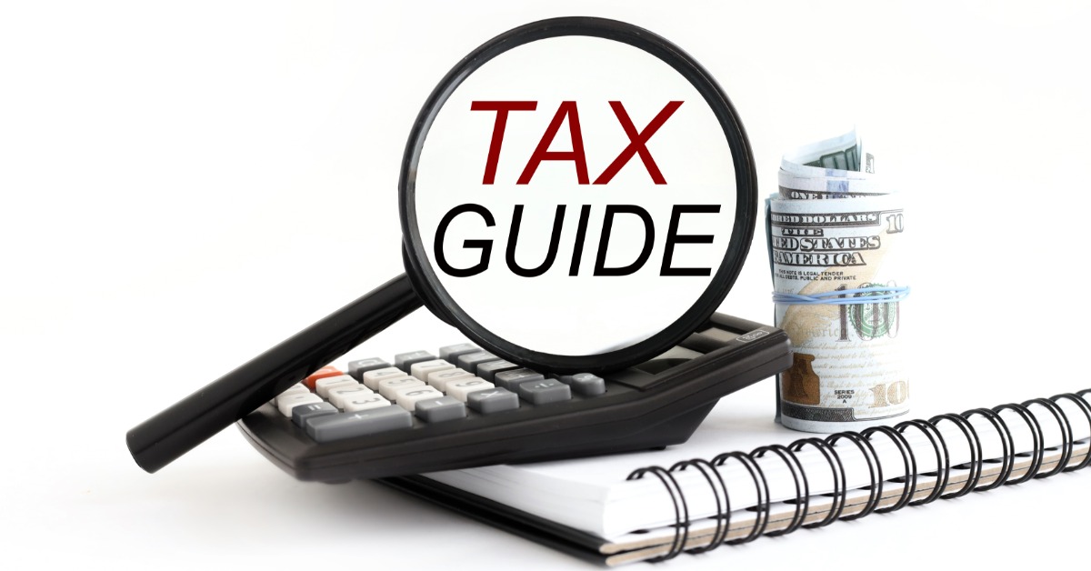 A magnifying glass that says "TAX GUIDE" in the spectacle on a calculator, notebook, and a roll of $100 bills.
