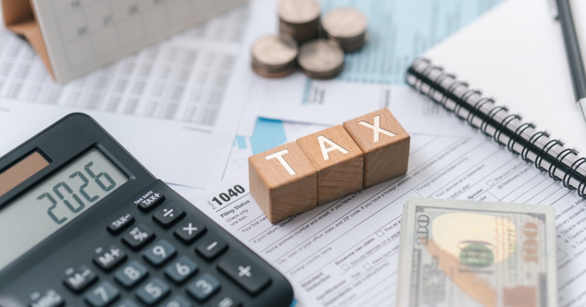 A close-up of three wooden letter blocks that spell out "TAX." Near the blocks are a calculator, dollar bills, and papers.
