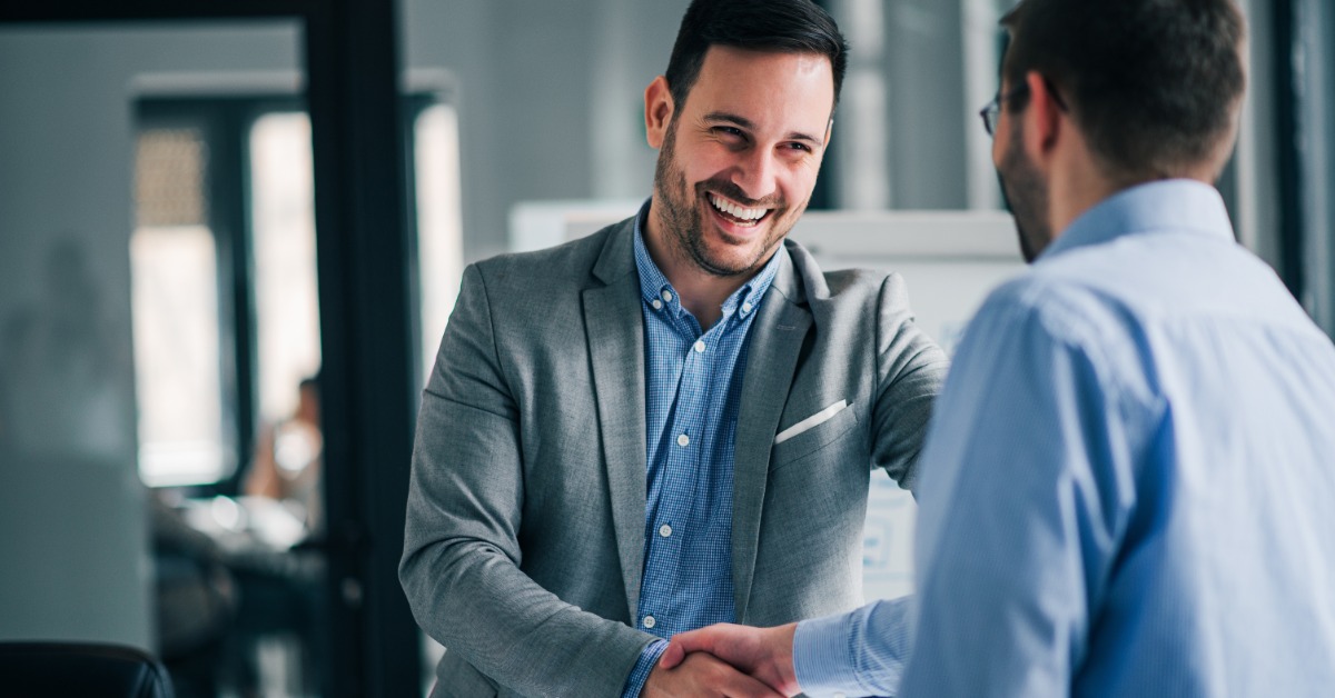 A man in a suit with black hair and a beard smiles while shaking the hands of another man in an office.