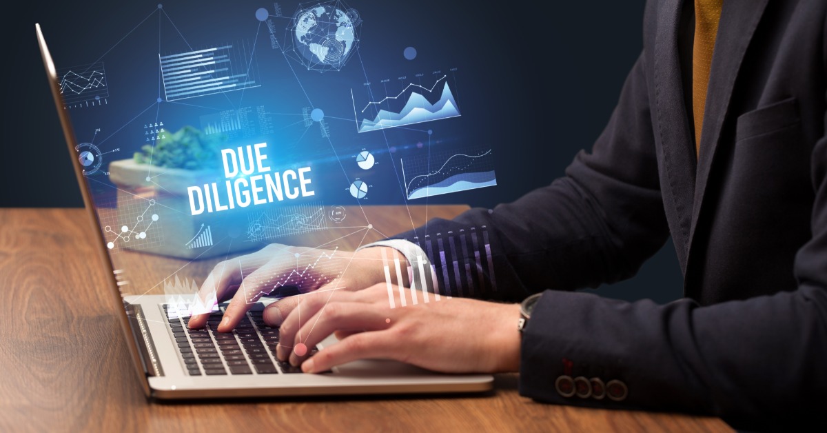 A close-up of a man in a suit typing on a laptop with a holographic above that reads "DUE DILIGENCE."