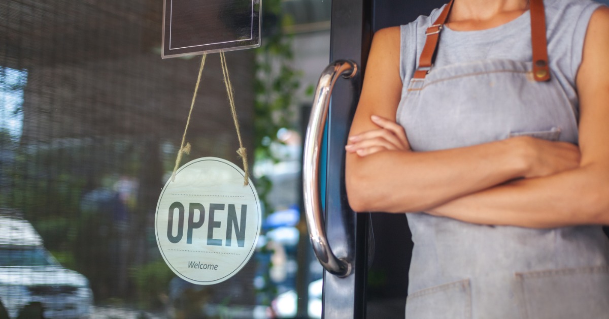 A woman in a grey apron leans against an open sliding door with a sign in the window that reads "OPEN" and "WELCOME."