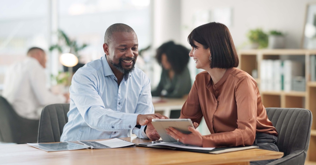A man and a woman in professional attire sitting at a table talking with electronic tablets and business documents.