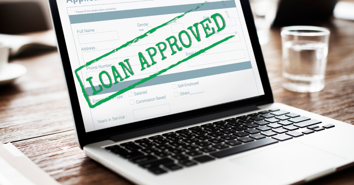 A close-up of a laptop on a table with an application form on the screen and "LOAN APPROVED" stamped across it.