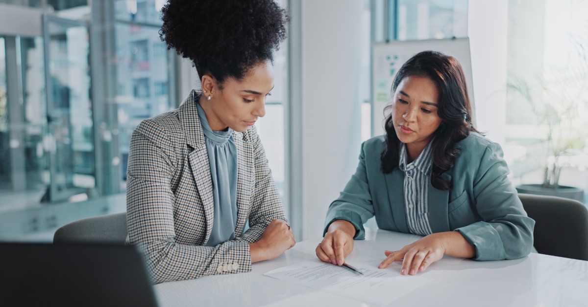 Two women in business attire are sitting at a table in a modern office, looking at paperwork together.